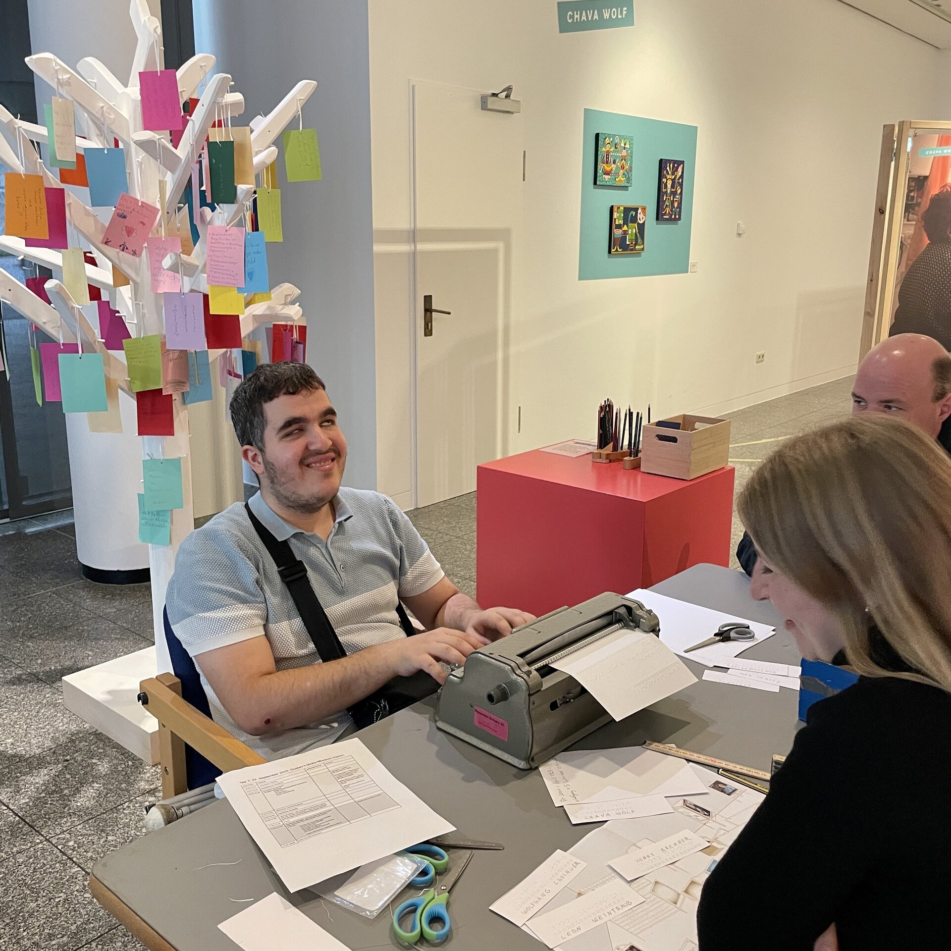 Personen sitzen an einem Tisch mit viel Papier. Eine Person schreibt an einer Schreibmaschine.